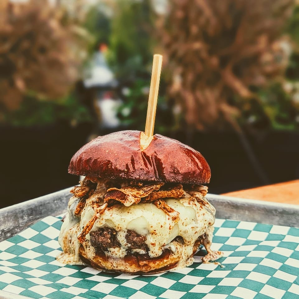 Stacked cheeseburger with a dark bun and skewer, served on a green-and-white checkered tray.