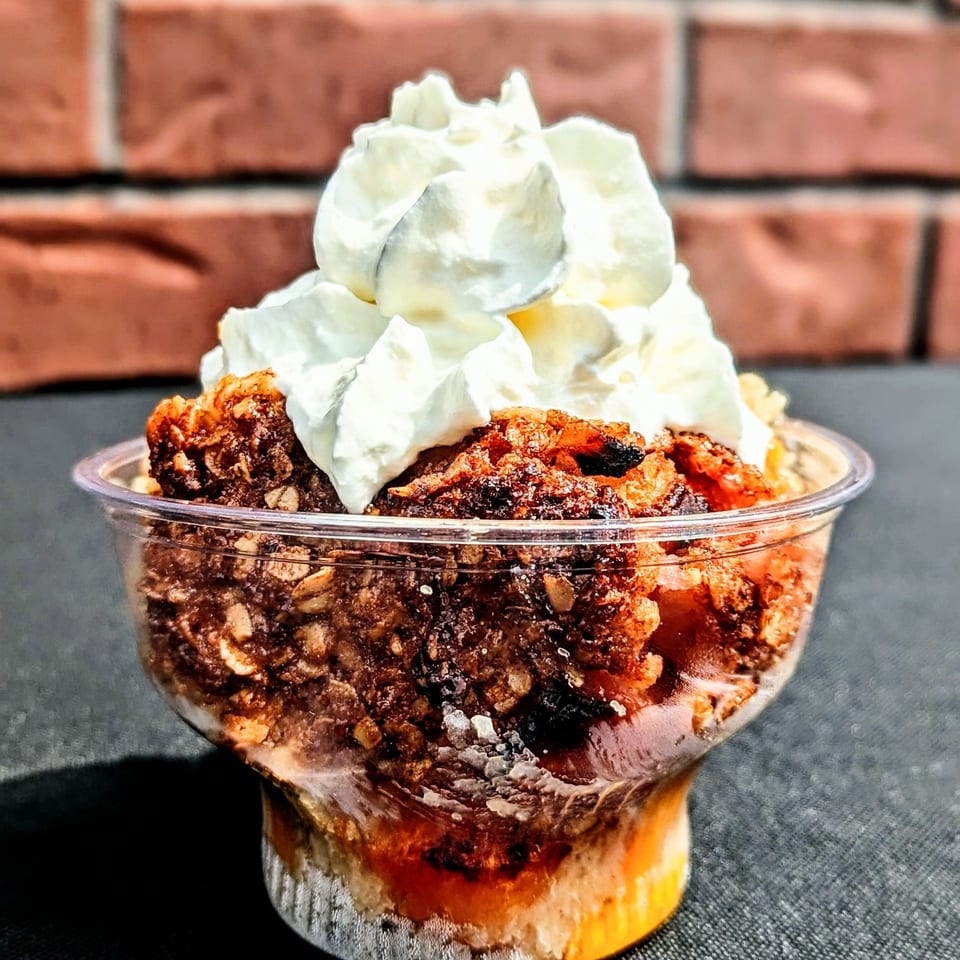 Layered dessert with chocolate crumble, caramel sauce, and whipped cream served in a clear cup against a brick wall.