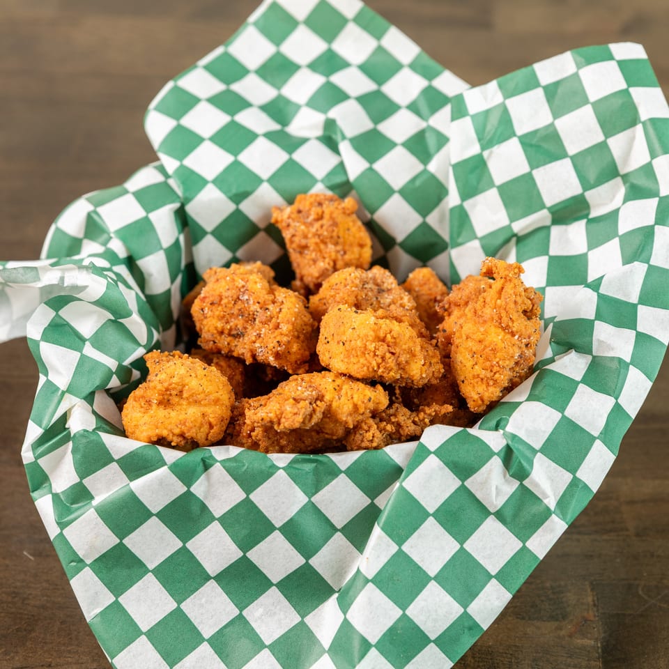 Basket of golden fried chicken pieces nestled in green-and-white checkered paper on a wooden table.