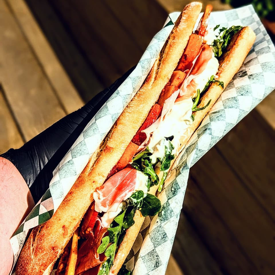 Loaded Italian sub sandwich with salami, ham, cheese, tomato, greens, and sauce in checkered paper, held outdoors.