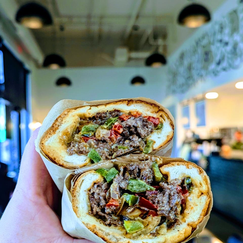 Hand holding two toasted flatbread sandwiches filled with grilled meat, green peppers, tomatoes, onions, and sauce.