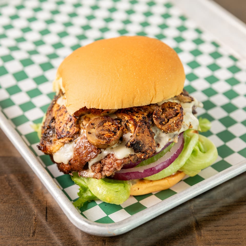 Close-up of a cheeseburger topped with grilled mushrooms, lettuce, and red onion on a green-and-white checkered tray.