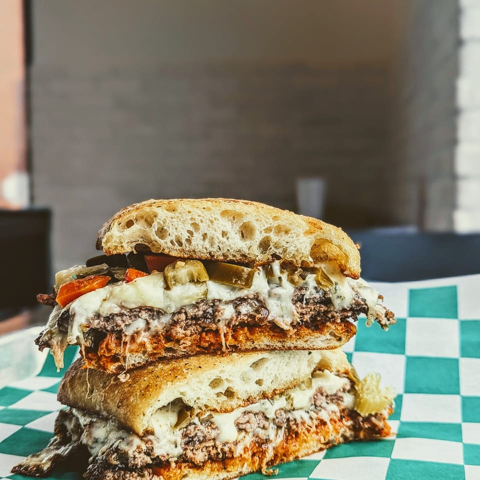 Towering double cheeseburger with pickles, carrots, crispy toppings, and creamy sauce on a green-and-white checkered tray.
